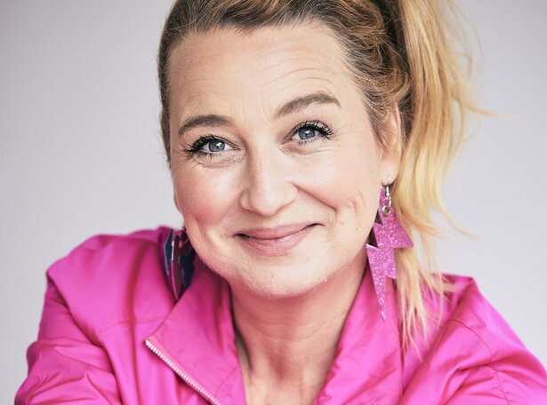 Frau mittleren Alters mit blondem Pferdeschwanz, pinker Jacke und auffälligen pinken Ohrringen in Blitzform, schaut belustigt in die Kamera.