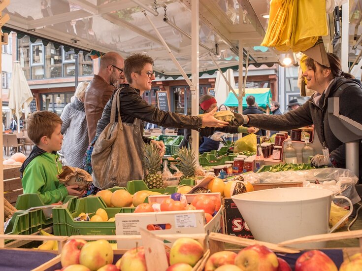 Eine lebhafte Szene auf dem Wochenmarkt in Bad Saulgau. Eine Frau in einer Lederjacke kauft Waren bei einer Verkäuferin und hält dabei eine wiederverwendbare Tasche in der Hand. Neben ihr steht ein Junge, der eine Papiertüte hält. Der Verkaufsstand ist gefüllt mit frischen Produkten wie Äpfeln, Ananas und anderem Obst. Die Verkäuferin lächelt, während sie die Waren überreicht. Im Hintergrund sind weitere Käufer und bunte Marktstände zu sehen.