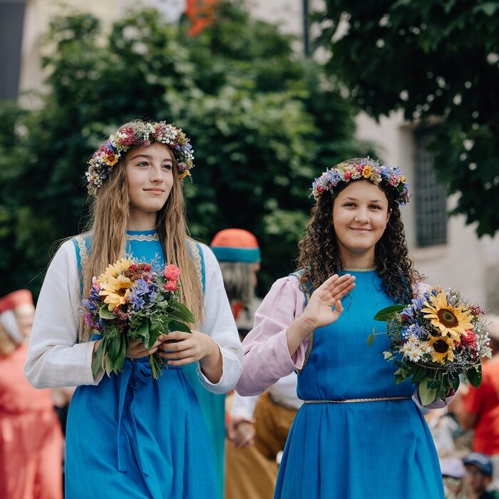 Frauen in Kleidern mit Blumenkränzen auf dem Kopf.