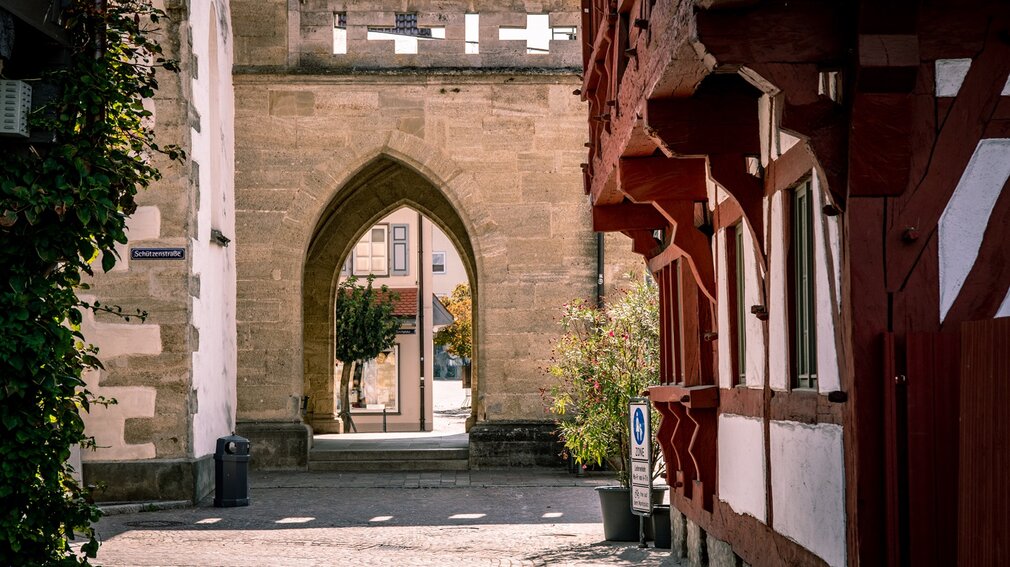 Stadtansicht eines historischen Kirchenportals mit Spitzbogen auf dem Marktplatz in Bad Saulgau, flankiert von Fachwerkhäusern und einer mit Pflanzen bewachsenen Mauer. Ein kleines Straßenschild mit der Aufschrift 'Schützenstraße' ist an der linken Wand angebracht, während im Hintergrund ein Baum und eine Häuserreihe zu sehen sind.