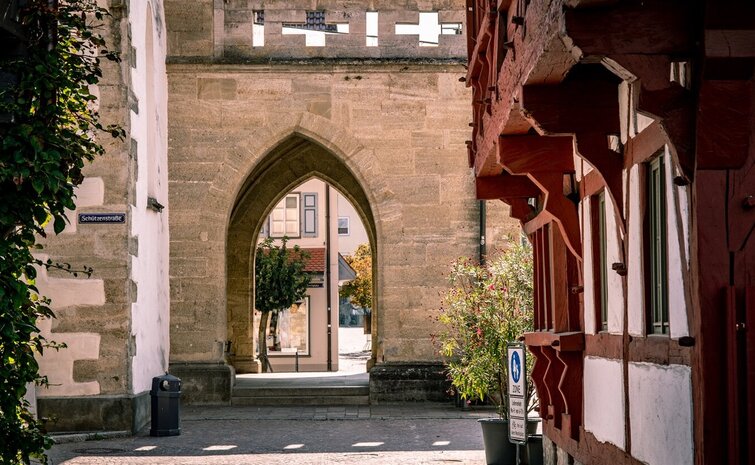 Stadtansicht eines historischen Kirchenportals mit Spitzbogen auf dem Marktplatz in Bad Saulgau, flankiert von Fachwerkhäusern und einer mit Pflanzen bewachsenen Mauer. Ein kleines Straßenschild mit der Aufschrift 'Schützenstraße' ist an der linken Wand angebracht, während im Hintergrund ein Baum und eine Häuserreihe zu sehen sind.