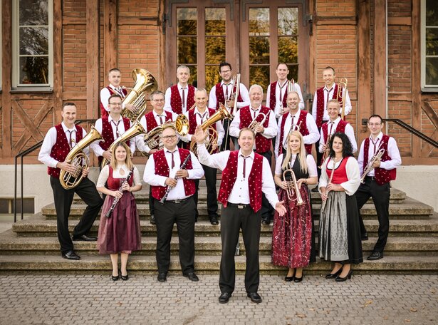Gruppe von Menschen in traditioneller Tracht mit Blasinstrumenten vor einem Backsteingebäude auf einer Treppe