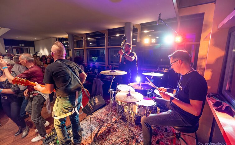 Liveband spielt vor dicht gedrängtem Publikum in einem hellen Veranstaltungsraum; im Vordergrund ein Schlagzeuger am Drumset, daneben Gitarrist und Kontrabassist, farbiges Bühnenlicht und tanzende Gäste sorgen für ausgelassene Konzertstimmung.