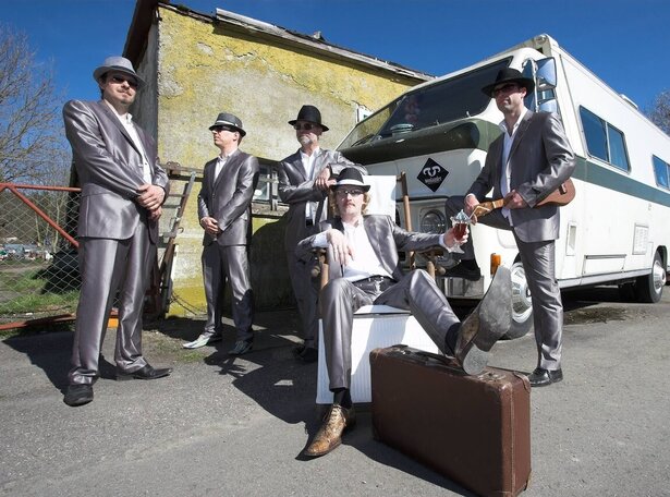Fünf Musiker in silbernen Anzügen und Hüten posieren lässig vor einem Oldtimer-Wohnmobil, ein Bandmitglied sitzt entspannt auf einem Stuhl mit Koffer im Vordergrund – urbaner Retro-Look mit Blues- und Rock’n’Roll-Flair.