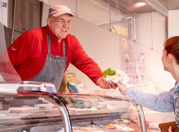 Markthändler in rot gestreifter Schürze reicht Kundin mit Jeansjacke eine Ware hinter einer Theke mit Schutzscheibe