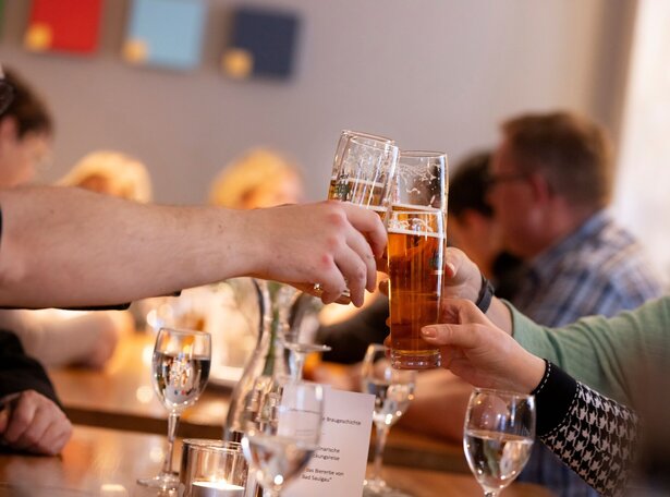 Zwei Personen stoßen mit Biergläsern an, im Hintergrund unscharfe Gäste an einem gedeckten Tisch