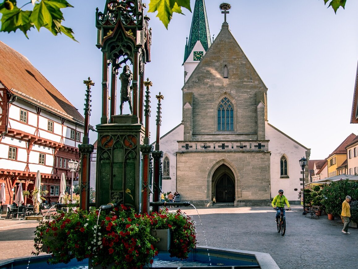 Historischer Marktplatz von Bad Saulgau mit gotischer Kirche, einem verzierten Brunnen mit Blumen sowie traditionellen und modernen Gebäuden; Menschen flanieren und ein Radfahrer fährt vorbei, bei sonnigem, Herbst-Wetter.
