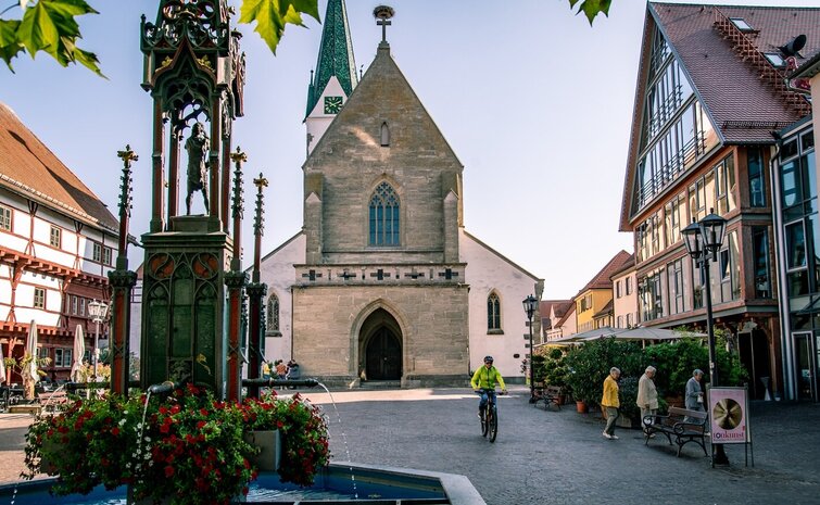 Historischer Marktplatz von Bad Saulgau mit gotischer Kirche, einem verzierten Brunnen mit Blumen sowie traditionellen und modernen Gebäuden; Menschen flanieren und ein Radfahrer fährt vorbei, bei sonnigem, Herbst-Wetter.