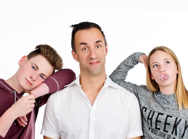 Mittelalter Mann in weißem Hemd, der teils genervt teils belustigt in die Kamera sieht, an seiner rechten Schulter lehnt ein Teenager, der genervt in die Kamera sieht, an der linken Schulte eine Teenagerin, die zur Seite sieht und eine Kaugummiblase macht.