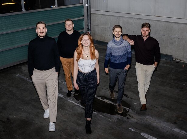 Bandfoto von fünf Musiker:innen, die selbstbewusst auf die Kamera zugehen; in der Mitte eine Frau mit rotem Haar, flankiert von vier Männern, aufgenommen in einer urbanen Industriehalle mit Betonboden und Metallwänden.