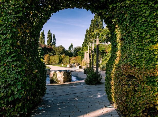 Blick durch einen von dichtem Grün bewachsenen Torbogen auf gepflasterten Weg mit großen Steinen und weiteren begrünten Strukturen im Kurgarten Bad Saulgau