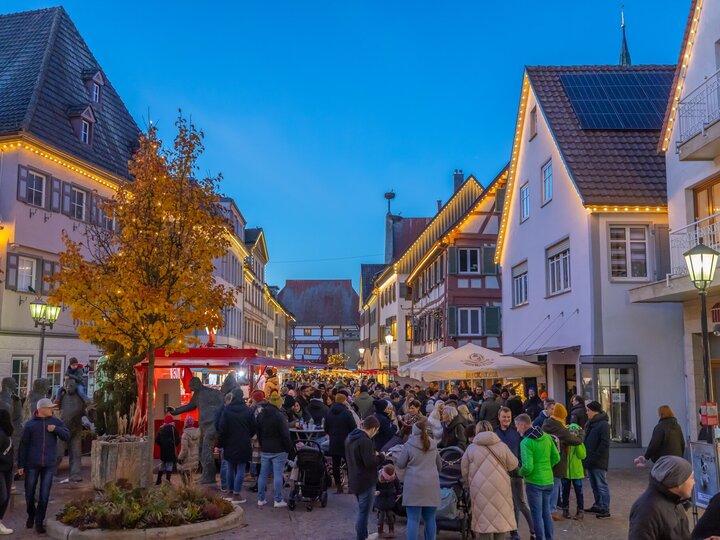 Abendliche Szene auf dem belebten Nikolausmarkt in Bad Saulgau. Menschenmengen schlendern durch eine mit Lichterketten geschmückte Straße, umgeben von Fachwerkhäusern und moderner Architektur. Ein roter Verkaufsstand und ein Baum mit herbstlich gelben Blättern stehen im Vordergrund.