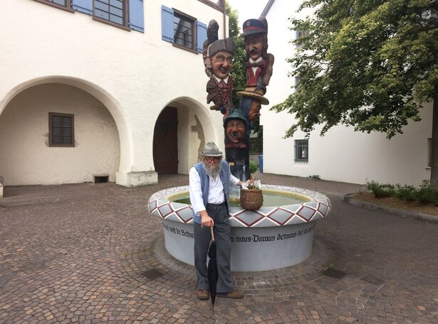 Älterer Mann mit Hut, Weste und Stock steht vor einem runden Brunnen mit bemaltem Rand und einer Säule mit mehreren bunten Figuren, umgeben von gepflastertem Platz und weißen Gebäuden mit Rundbögen