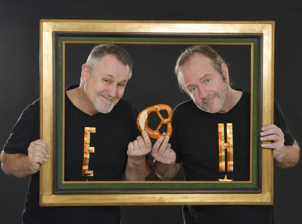 Zwei Männer halten gemeinsam einen goldenen Bilderrahmen und präsentieren darin eine Brezel, beide lächeln in die Kamera vor dunklem Hintergrund