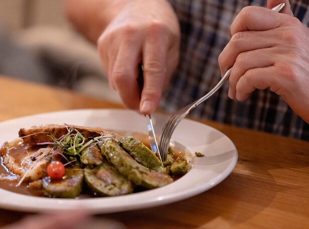 Nahaufnahme von Händen, die mit Messer und Gabel grüne, gerollte Speisen auf einem weißen Teller mit Soße und kleinen Tomaten schneiden