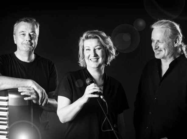 Schwarz-weißes Bandporträt von drei Musiker:innen: eine Frau in der Mitte hält ein Mikrofon und lächelt in die Kamera, links steht ein Mann mit einem Akkordeon, rechts ein weiterer Mann blickt lächelnd zur Sängerin; dunkler Hintergrund.