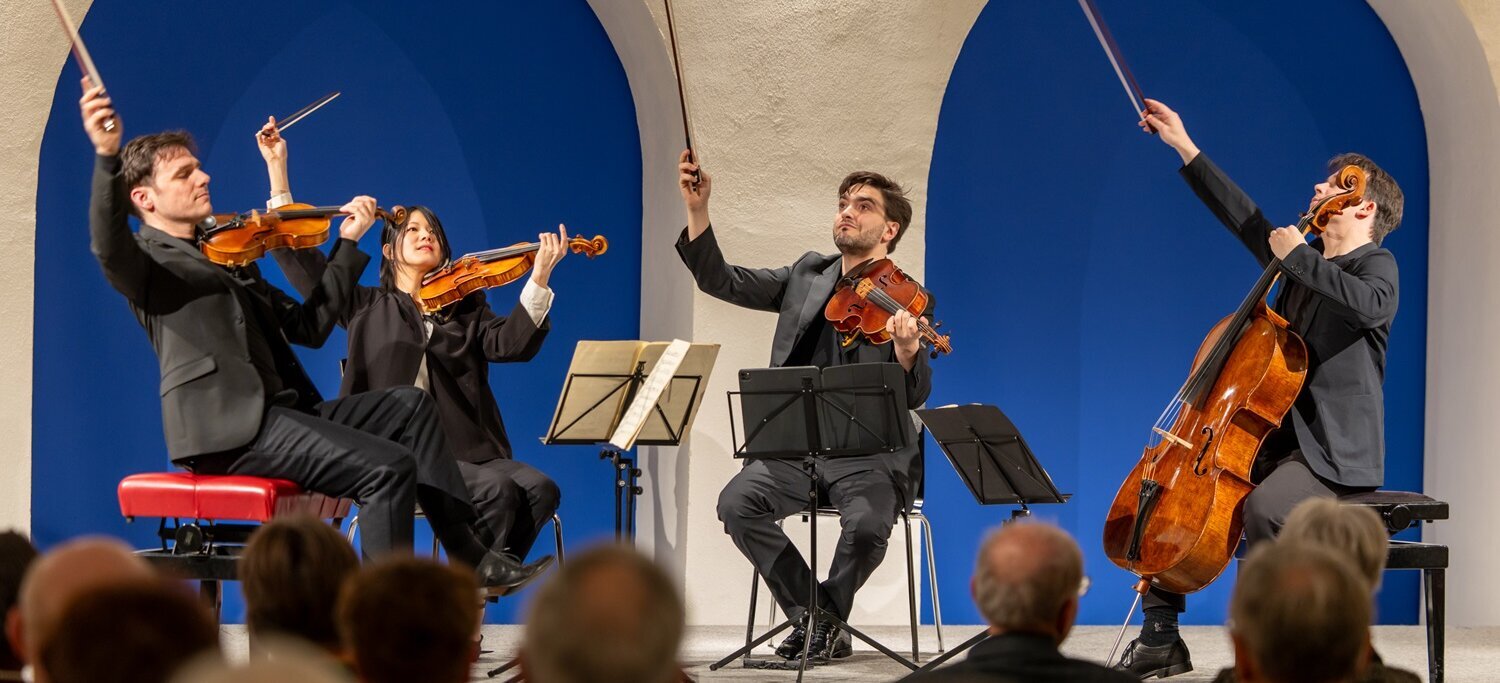 Streichquartett mit zwei Geigern, einem Bratschisten und einem Cellisten spielt vor Publikum in einem Raum mit weißen Rundbögen vor blauer Wand.