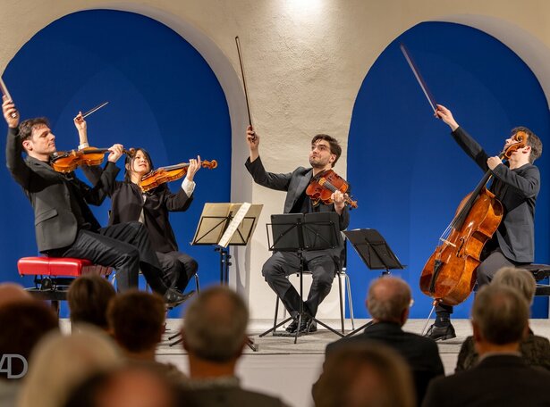 Streichquartett mit zwei Geigern, einem Bratschisten und einem Cellisten spielt vor Publikum in einem Raum mit weißen Rundbögen vor blauer Wand.