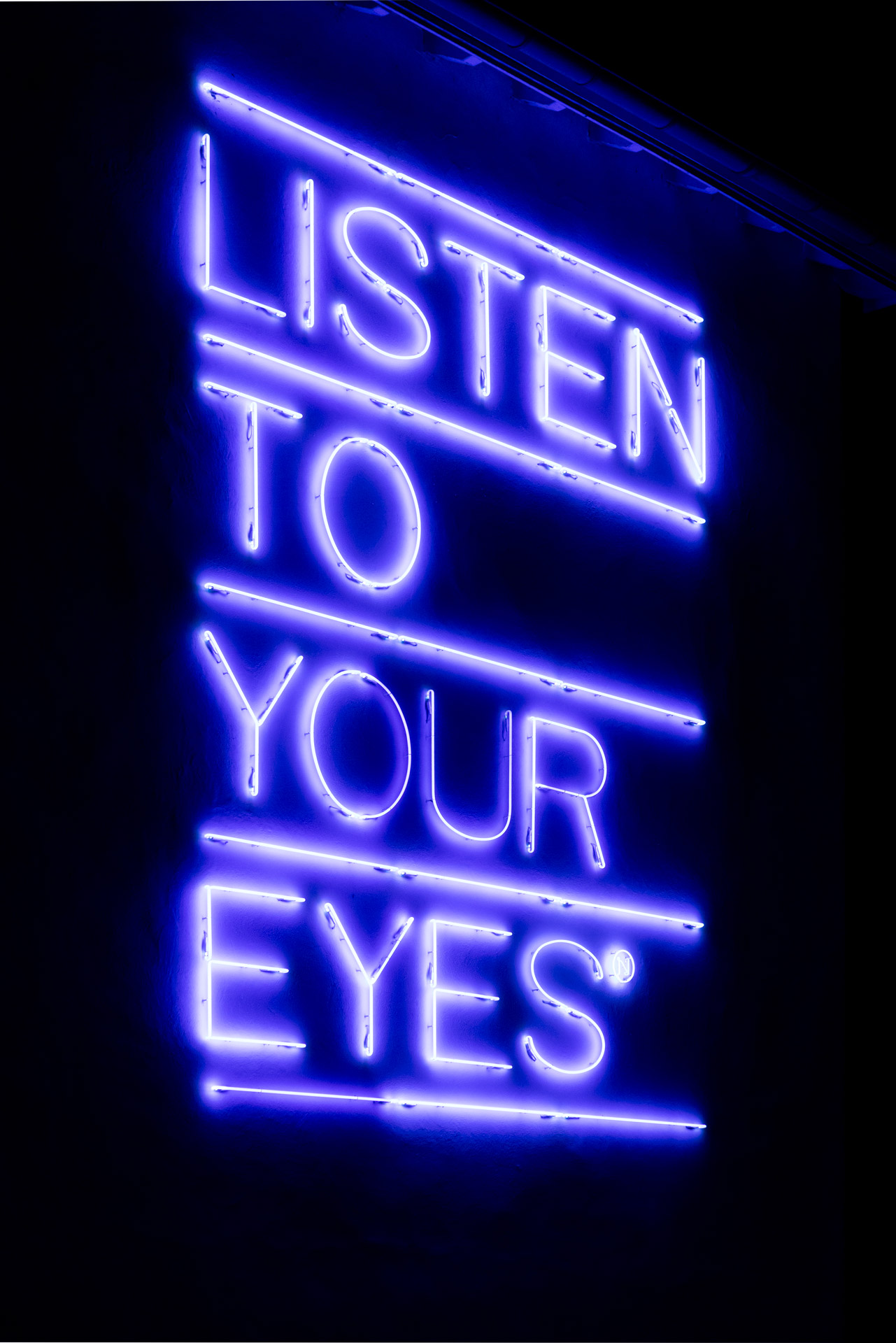 Leuchtendes Neonschild mit dem Text 'LISTEN TO YOUR EYES' in Großbuchstaben und Unterstreichungen des Künstlers Maurizio Nannucci